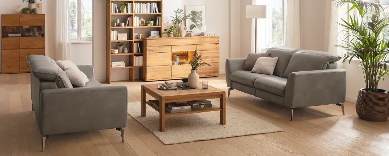Modernes Wohnzimmer mit zwei Sofas, Couchtisch und Sideboard aus Massivholz. Helle Wohnmöbel mit Regal, Pflanzen und Teppich schaffen ein natürliches Wohnkonzept für stilvolles und nachhaltiges Wohnen.